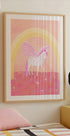 Ein gerahmtes Poster eines Einhorns mit einem rosa-weißen Farbschema vor einem rosa-gelben Hintergrund mit einem Regenbogenbogen.